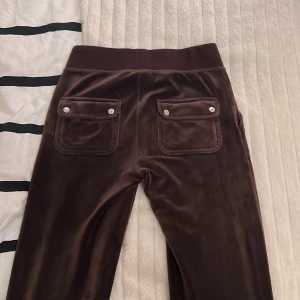 Bruna mjukisbyxor från Juicy Couture - Snygga bruna mjukisbyxor från Juicy Couture i en mjuk och skön velour. Byxorna har två bakfickor med knappar och en broderad logga på ena benet. Perfekta för en avslappnad stil. Har använt fyra gånger, köpte för 1200