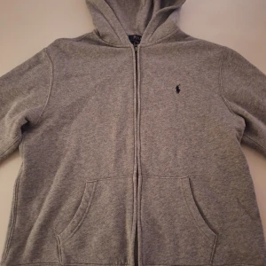 Grå hoodie från Ralph Lauren - Säljer en stilren grå hoodie från Ralph Lauren med dragkedja och klassisk logga på bröstet. Perfekt för en avslappnad look. Den har en bekväm passform och praktiska fickor framtill.