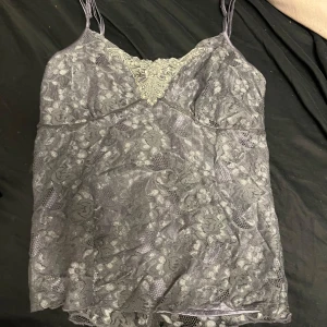 Grå spetslinne - Säljer ett elegant grått spetslinne med smala axelband och v-ringning. Perfekt för en stilren look med vackra spetsdetaljer som ger en romantisk känsla.