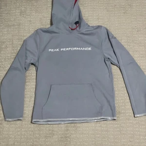 Grå hoodie från Peak Performance - Säljer en stilren grå hoodie från Peak Performance med vit logga på bröstet. Tröjan har en praktisk känguruficka och justerbar huva med röd insida och dragsko. Perfekt för kyliga dagar.