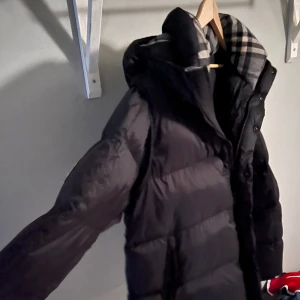 Svart dunjacka från Burberry med avtagbara ärmar - Säljer en snygg svart dunjacka från Burberry med klassiskt rutigt foder. Jackan har en dragkedja och knappar framtill samt en hög krage för extra värme. Perfekt för kyliga dagar! Man kan även ta av ärmarna och ha den som en väst under våren. (ÄKTA!!) Jackan är som ny inga skador eller liknande