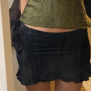 Svart mini kortkjol med volangdetaljer - Säljer en superfin svart kortkjol med volangdetaljer. Perfekt för både fest och vardag! Kjolen har en omlott-look och knytning i sidan som ger en snygg passform. Den är i bra skick och passar perfekt till en cool topp eller blus.