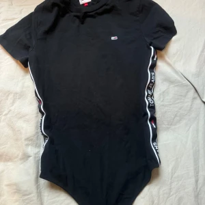 Svart klänning från Tommy Jeans - Snygg svart kjol och topp från Tommy Jeans med korta ärmar och logotypband längs sidorna.  Använd 1 gång Kjolen har storlek M och Toppen storlek S