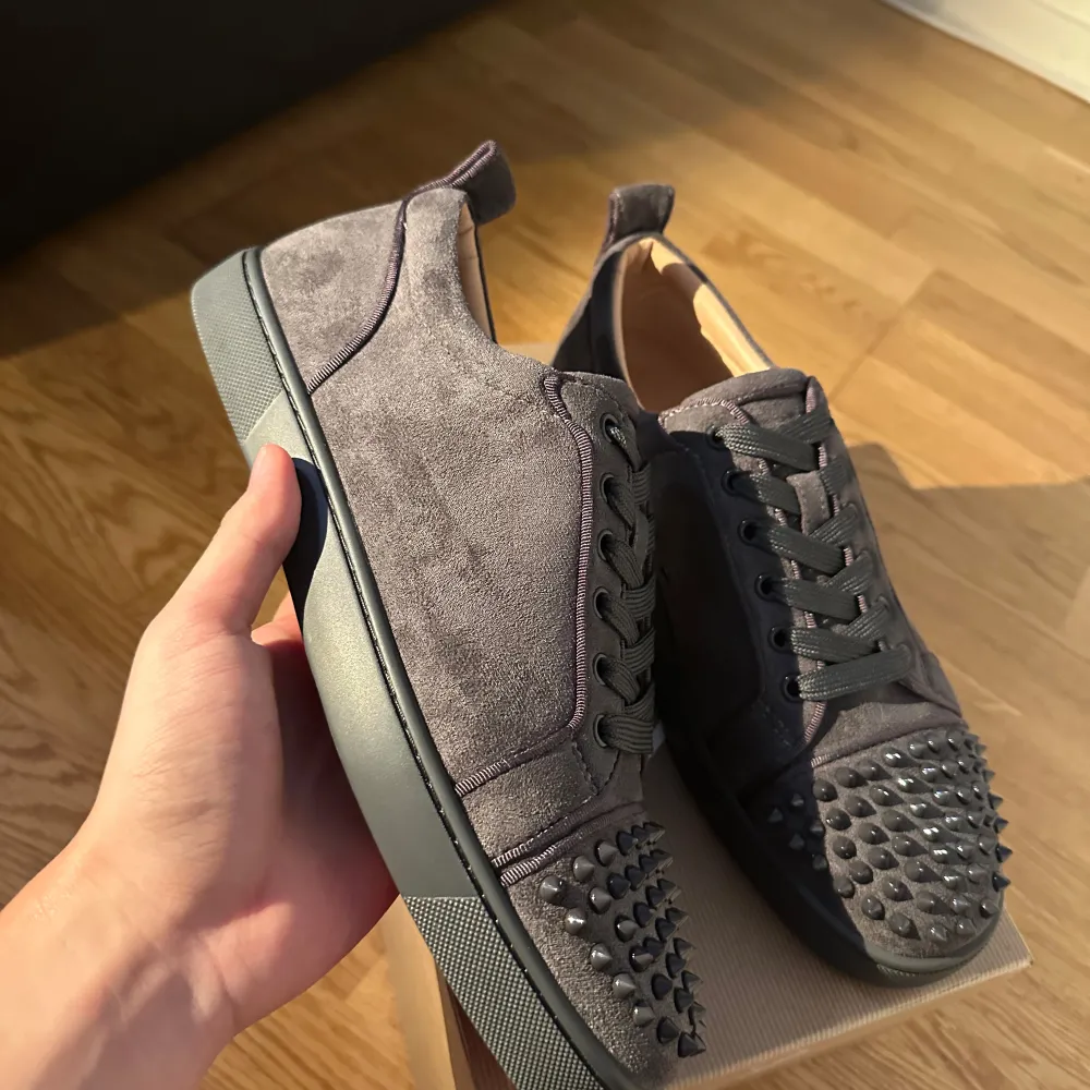 Säljer ett par grå sneakers i mocka från Louboutin. De har en cool design med nitar på tån och snörning. Sulan är svart med den ikoniska röda undersidan. Perfekta för att ge din outfit en edgy touch!. Skor.