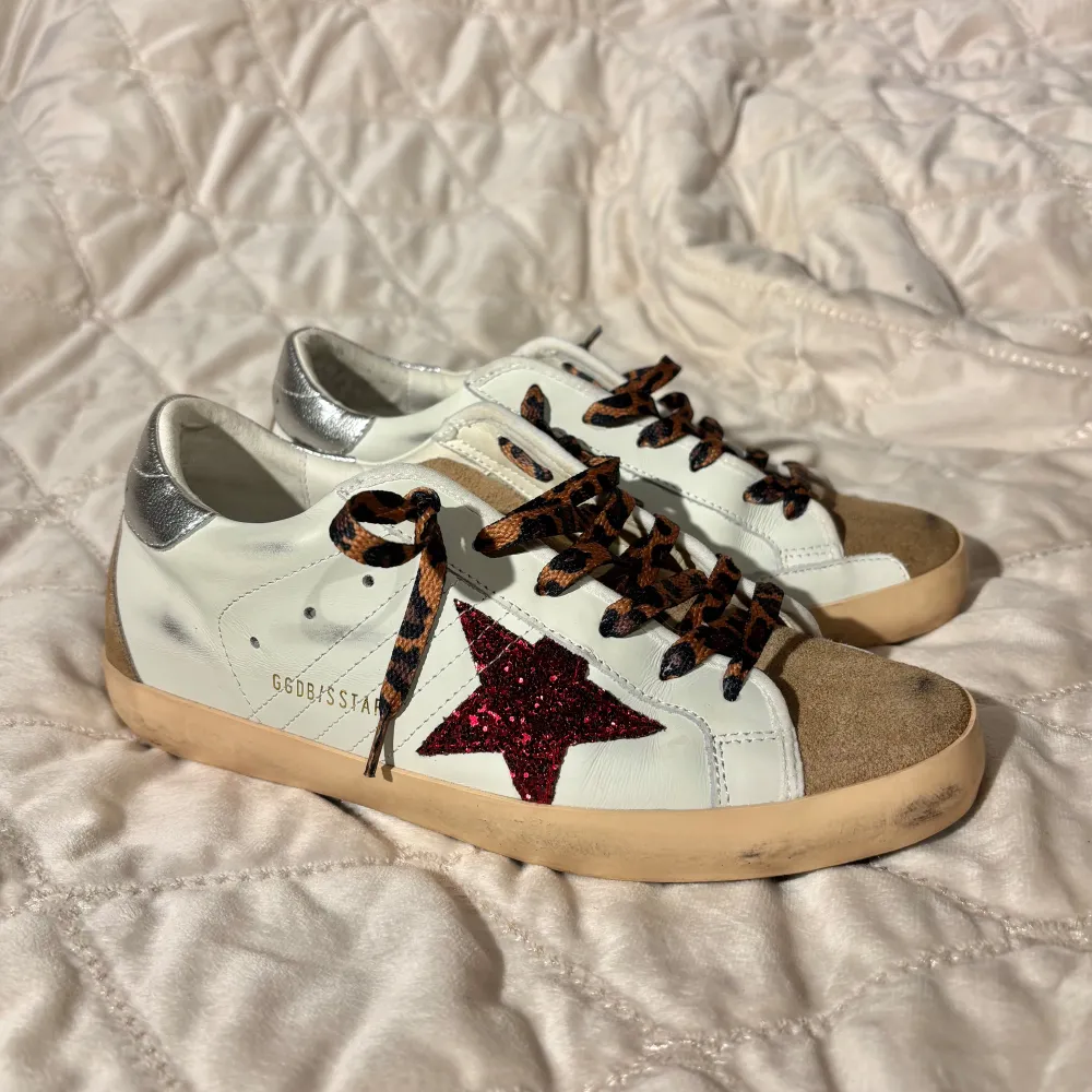 Säljer ett par Golden Goose sneakers i vitt skinn med en röd glittrig stjärna på sidan. Skorna har leopardmönstrade snören och silverdetaljer på hälen. De är i mycket bra skick och är endast använda ett fåtal gånger då dom är för små för mig. De är storlek 38 men skulle säga de även kan passa dig som har storlek 37. Pris går att diskutera vid snabba affärer!. Skor.