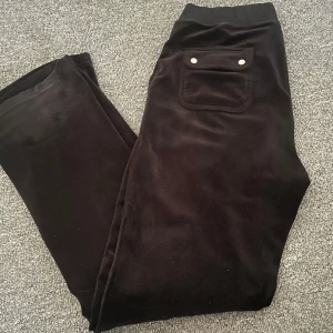 Svarta mjukisbyxor från Juicy Couture - Säljer ett par svarta mjukisbyxor från Juicy Couture i storlek M. Byxorna har en bekväm passform med elastisk midja och dekorativa knappar på fickan. Perfekta för en avslappnad dag hemma eller en promenad i parken.