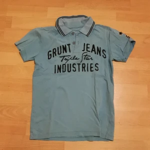 Ljusblå pikétröja från Grunt Jeans - Säljer en ljusblå pikétröja från Grunt Jeans med tryck på bröstet och en cool stjärndetalj på ärmen. Tröjan har en klassisk krage med knappar och svarta ränder. Perfekt för en avslappnad stil! Står M på tröjan, men skulle ändå säga att den mer passar S! 