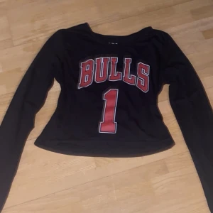 Svart långärmad Bulls tröja från BNDS - Cool svart långärmad tröja från BNDS med 'BULLS' och siffran '1' i rött på framsidan. Perfekt för en avslappnad stil eller sportiga tillfällen. Tröjan är i ett mjukt material och har en croppad passform. Passar perfekt till jeans eller kjol för en trendig look. Tröjan är one size men skulle säga den passar allt från XS-tajt M