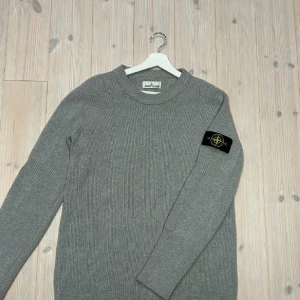 Grå stickad tröja från Stone Island - Säljer en snygg grå stickad tröja från Stone Island. Den har en klassisk design med ribbad struktur och en ikonisk logga på ärmen. Perfekt för både höst och vinter, och den är i mycket bra skick. Passar bra till både vardag och lite finare tillfällen. Skriv vid minsta fråga😊