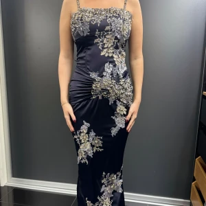 Mörkblå långklänning med blommig spets - Elegant mörkblå långklänning i satin med smala axelband och vacker blommig spetsdetalj. Perfekt för festliga tillfällen med sin figurnära passform och stilrena design. Klänningen har en dragkedja i ryggen för enkel påklädning.