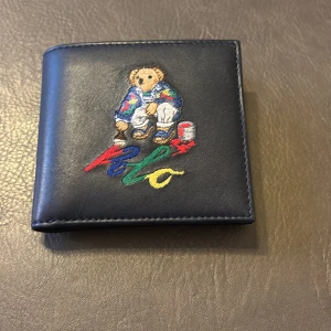 Marinblå plånbok från Polo Ralph Lauren - Säljer en mörkblå unik plånbok från Polo Ralph Lauren. Den har ett broderat motiv av en nalle på framsidan. Plånboken har flera fack för kort, kontanter och en myntficka. Matrialet är i Buffalo läder. Använd 1 gång. Nypris 2500 kr. Originalbox medföljer. Priset går att diskutera.