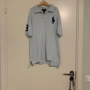 Ljusblå pikétröja från Ralph Lauren - Säljer en snygg ljusblå pikétröja från Ralph Lauren. Den har en klassisk krage och korta ärmar med en stor mörkblå logga på bröstet och siffran 3 på ärmen. Perfekt för en avslappnad men stilren look. Passar både till vardags och fest!
