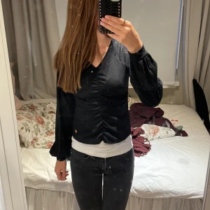 Svart ribbad topp med v-ringning - Säljer en snygg svart ribbad topp med v-ringning och långa ärmar. Toppen har en smickrande rynkad detalj framtill som ger en fin passform. Perfekt för både vardag och fest! 🖤