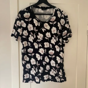 Svart blommig topp från J.Lindeberg - Säljer en snygg svart tshirt från J.Lindeberg med ett blommönster i vitt. Tshirten har korta ärmar och en rund halsringning. Perfekt för både vardag och fest!