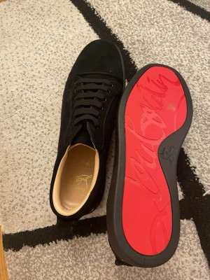Svarta sneakers med röd sula - Snygga svarta sneakers från Christian Louboutin med klassisk röd sula och vit broderad logga på hälen. Skorna har en låg profil, svarta skosnören och ovandel i mocka. Perfekta för dig som vill sticka ut med exklusiv design.