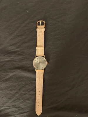 Beige klocka med silverfärgad urtavla - Stilren klocka med beige läderband och rund, silverfärgad urtavla. Urtavlan har minimalistiska svarta markeringar och guldfärgade detaljer. Perfekt accessoar för dig som gillar enkel och klassisk design.