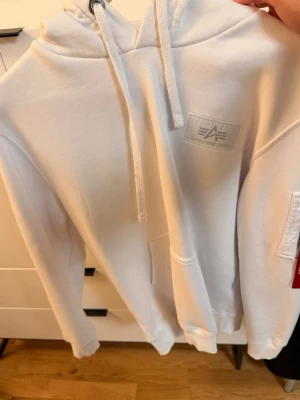 Vit hoodie från Alpha Industries - Snygg vit hoodie från Alpha Industries med stor svart logga på ryggen och mindre logga på bröstet. Hoodien har huva med dragsko och en klassisk känguruficka framtill. Perfekt för en avslappnad och trendig stil.