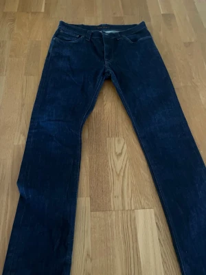 Nudie jeans  - Säljer ett par mörkblå jeans med rak passform och klassisk femficksdesign. Jeansen har kontrastsömmar och detaljer på bakfickorna. Tillverkade i denim med en stilren look som passar till det mesta. Nudie jeans storlek w33 L34