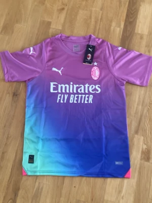 AC Milan x Puma färgglad matchtröja - Säljer en AC Milan matchtröja från Puma i en snygg färgskiftning från lila till blå och turkos. Tröjan har korta ärmar, rund hals och tryckta loggor samt sponsortryck på både fram- och baksida. Perfekt för fotbollsträning eller att bära som supporter.