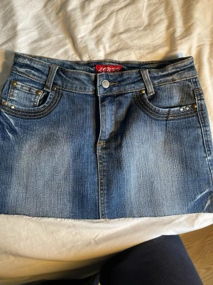 Blå jeanskjol  - Snygg blå jeanskjol i kort modell med slitna detaljer och dekorativa nitar på fickorna. Den är low waist och lagom kort. Säljer den pågrund av att den aldrig kommer till användning!😊💓