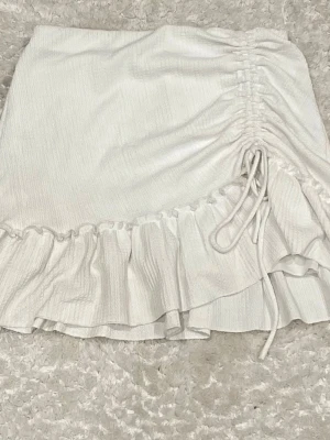 Vit kjol med volang och snörning - Supersöt vit kjol från Gina Tricot med volangkant och snörning på sidan som ger en snygg rynkad effekt. Kjolen är kort och har en lätt struktur i tyget, perfekt för en trendig look. Tillverkad i polyesterblandning.