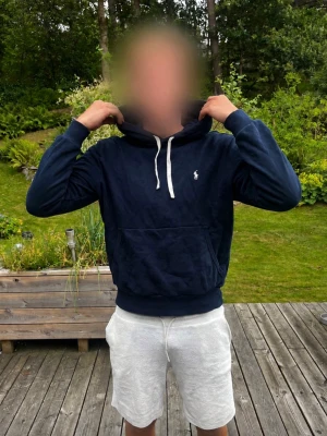 Mörkblå hoodie med huva från Polo Ralph Lauren - Snygg mörkblå hoodie från Polo Ralph Lauren med klassisk vit logga på bröstet. Hoodien har huva med vita dragsnören och en stor magficka framtill. Tillverkad i mjukt bomullsmaterial och har ribbade muddar vid ärmslut och nederkant.