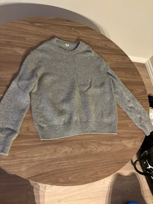 Grå stickad tröja med rund hals från Arket - Mysig grå stickad tröja med rund hals och ribbade muddar vid ärmslut och nederkant. Perfekt för lager på lager och enkel att matcha. Tröjan har en avslappnad passform och är tillverkad i mjukt material.
