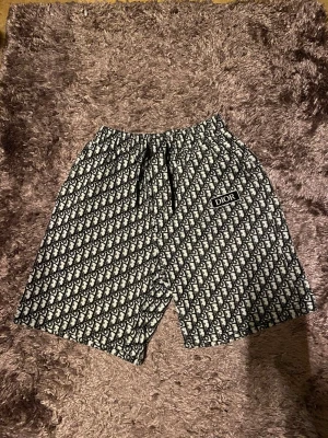 Di*r shorts - Säljer ett par svarta och vita shorts från med ett all-over monogrammönster och snörning i midjan. Shortsen har en tydlig logga på ena benet och är perfekta för dig som vill sticka ut med en lyxig streetwear-look.