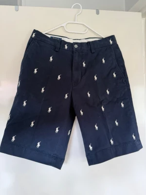 Polo Ralph Lauren Shorts – All over pony embroidery – Marinblå – Storlek W30 - Snygga mörkblå shorts med vita broderade loggor över hela plagget. Klassisk passform med bälteshällor, knapp och dragkedja framtill samt två bakfickor med knappar. Perfekta för sommaren och ger en stilren look. Storlek: Storlek 30 (S/M i midjan) 