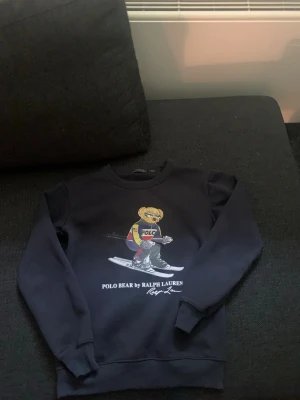 Svart sweatshirt Polo Bear Ralph Lauren - Cool svart sweatshirt från Ralph Lauren med den ikoniska Polo Bear-trycket framtill där björnen åker skidor. Tröjan har rund hals, långa ärmar och ribbade muddar. 