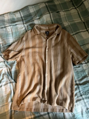 Beige randig skjorta med kort ärm - Säljer en beige skjorta med vita vertikala ränder och korta ärmar. Skjortan har klassisk krage och knäppning framtill. Tillverkad i ett lätt och luftigt material, perfekt för varmare dagar.