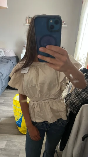 Beige blus med volang från LMTD - Supersöt beige blus från LMTD med volangärmar och markerad midja. Blusen har dekorativ spetsdetalj runt midjan och knäppning med pärlknapp i nacken. Tillverkad i ett glansigt, lätt material som ger en lyxig känsla. Tröjan har aldrig används.