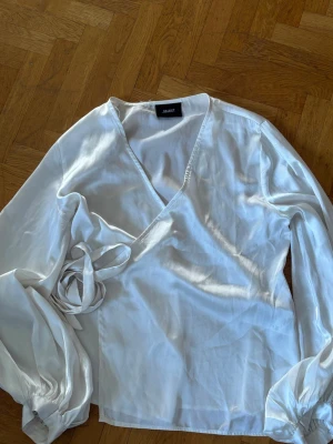 Vit omlottblus från Object - Snygg vit omlottblus från Object med långa ärmar och breda manschetter. Blusen har knytband i midjan och är tillverkad i ett glansigt, tunt material som ger en elegant look. Perfekt till jeans eller kjol för en trendig outfit.