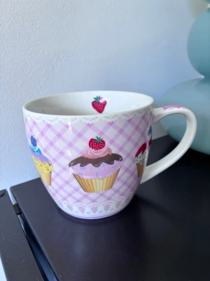 Söt mugg med cupcakes och jordgubbar - En stor, vit keramikmugg med lila rutigt mönster och färgglada cupcakes med bärmotiv runtom. Insidan har en liten jordgubbe som detalj. Perfekt för te eller varm choklad och ger en gullig touch till köket.