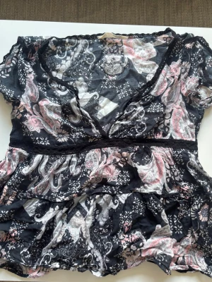 Svart blommig blus med spetsdetaljer - Snygg svart blus med blommigt mönster i vitt och rosa. V-ringad med spetsdetaljer längs halsringningen och midjan. Lätt och luftig modell med korta ärmar och volang nertill. Perfekt för dig som gillar romantisk och feminin stil.