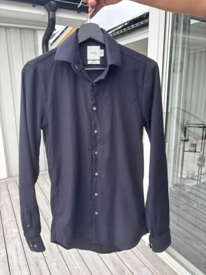 Calvin Klein Mörkblå skjorta med diskret mönster, strl S - Snygg mörkblå skjorta med diskret småmönster och klassisk krage. Skjortan har långa ärmar och knäppning framtill med vita knappar. Perfekt för dig som gillar stilrena plagg med en twist.