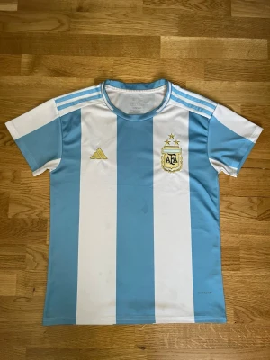 Argentina fotbollströja Adidas - Säljer en klassisk Argentina fotbollströja från Adidas med ljusblå och vita vertikala ränder. Tröjan har korta ärmar, rund hals och tre ljusblå ränder på axlarna. AFA-logga och Adidas-märke på bröstet. Perfekt för match eller träning.