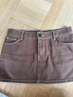 Brun kort jeanskjol med kontrastsömmar - Snygg brun kort jeanskjol med vita kontrastsömmar och klassiska fickor både fram och bak. Kjolen har bälteshällor och stängs med knapp och dragkedja framtill. Perfekt för dig som gillar en enkel men trendig stil.
