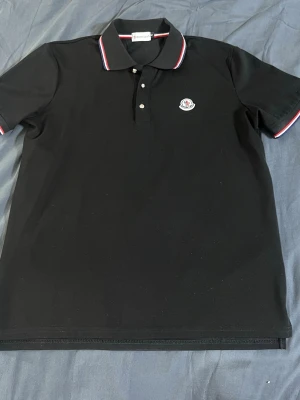 Svart pikétröja från Moncler - Klassisk svart pikétröja från Moncler med vit och röd rand på krage och ärmslut. Tröjan har knappslå framtill och Moncler-logga broderad på bröstet. Perfekt till en clean och stilren look.
