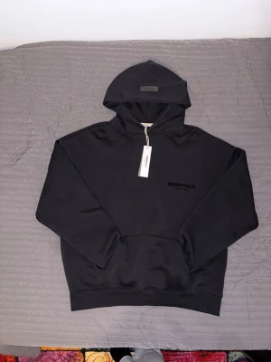 Svart Essentials set - Säljer en svart hoodie från Essentials  med diskret logga på bröstet och stor huva. Hoodien har en klassisk känguruficka framtill och ribbade muddar vid ärmslut och nederkant. Perfekt för en clean och stilren streetwear-look.
