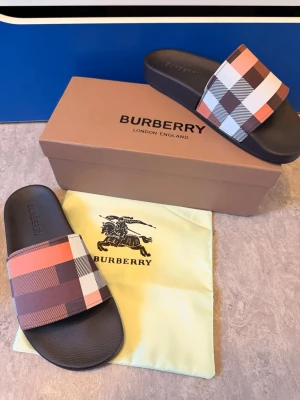 Burberry rutiga slides i svart och beige - Snygga slides från Burberry med klassiskt rutigt mönster i beige, brun, svart, vit och orange. Tofflorna har en bred rem över foten och svart sula i gummi. Perfekta för chill dagar eller sommarhäng. Kommer med originalkartong och dustbag.