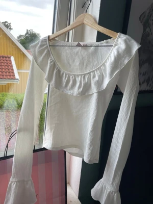 Vit långärmad topp med volang - Söt vit långärmad topp med bred volang runt ringningen och volangdetaljer vid ärmsluten. Aldrig använd, säljer då den endast tar plats i garderoben.
