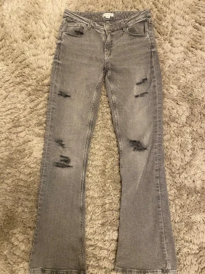 Grå slitna bootcut jeans - Säljer ett par grå jeans med bootcut-modell och slitna detaljer på benen. Jeansen har klassisk femficksdesign och råa kanter vid bensluten. Perfekta för en avslappnad och trendig look.