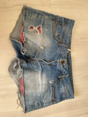 Ljusa slitna jeansshorts - Säljer ett par ljusblå jeansshorts med råa kanter och flera slitningar framtill. Shortsen har klassiska fem fickor, knappgylf och synliga sömmar i kontrastfärg. Perfekta för en avslappnad och trendig sommarstil.