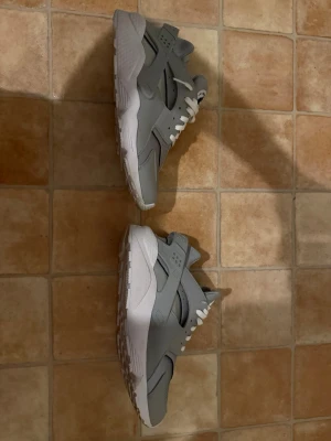 Nike Air Huarache grå sneakers - Snygga Nike Air Huarache sneakers i olika nyanser av grått med vita detaljer och vita skosnören. Skorna har en sportig design med perforerad tåbox och en stabil sula. Perfekta för dig som gillar streetwear och vill ha något bekvämt och stilrent.