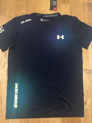 Svart tränings-tshirt Under Armour - Svart tränings-tshirt från Under Armour med reflekterande logga på bröstet och text på ärm och nederkant. Tillverkad i ett lätt och snabbtorkande material som passar perfekt för löpning eller gym. Klassisk passform med rund hals och korta ärmar.