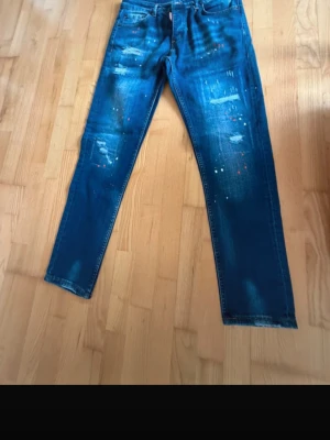 Blå distressed jeans från Dsquared2 - Säljer ett par blå jeans från Dsquared2 med slitningar, färgstänk och en cool, tvättad look. Jeansen har klassisk femficksmodell och orange kontrastsömmar. Perfekta för dig som gillar streetstyle och vill sticka ut lite extra.