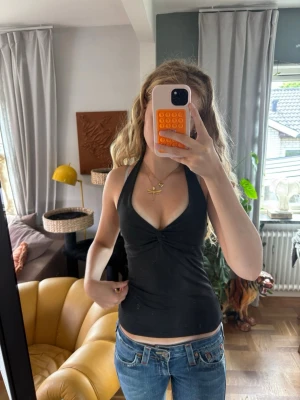 Svart halterneck linnetopp - Snygg svart halterneck linnetopp med v-ringning och twistad detalj framtill. Toppen är ärmlös och har en slim passform som framhäver figuren. Perfekt för varma dagar och enkel att matcha med jeans eller kjol.
