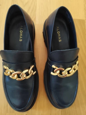 Svarta loafers med guldkedja från Alohas - Stilrena svarta loafers från Alohas i slätt läder med chunky guldfärgad kedjedetalj över vristen. Klassisk form med rundad tå och låg sula. Perfekta för dig som vill ha en trendig och elegant look med en twist.