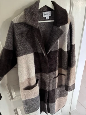 Randig ullkappa från Joseph Ribkoff - Snygg och mysig kappa från Joseph Ribkoff i mjuk ullmix. Kappan har breda blockränder i brunt, beige, grått och offwhite, stora slag och två fickor framtill. Perfekt för dig som vill ha en statement piece till höst och vinter.